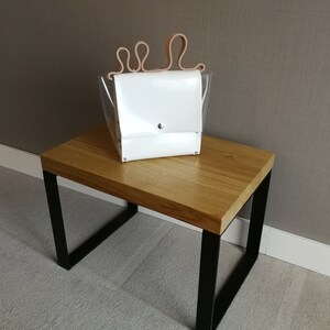 May include: A white leather handbag with a clear plastic body and a unique brown leather handle. The bag is sitting on a wooden table with black metal legs.