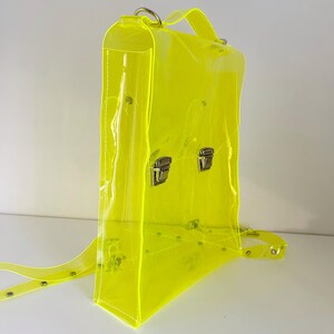 May include: A neon yellow transparent backpack with silver-colored metal clasps and hardware. The bag has adjustable straps and a top handle. The clear material allows visibility of the contents inside. The backpack is designed with a rectangular shape.