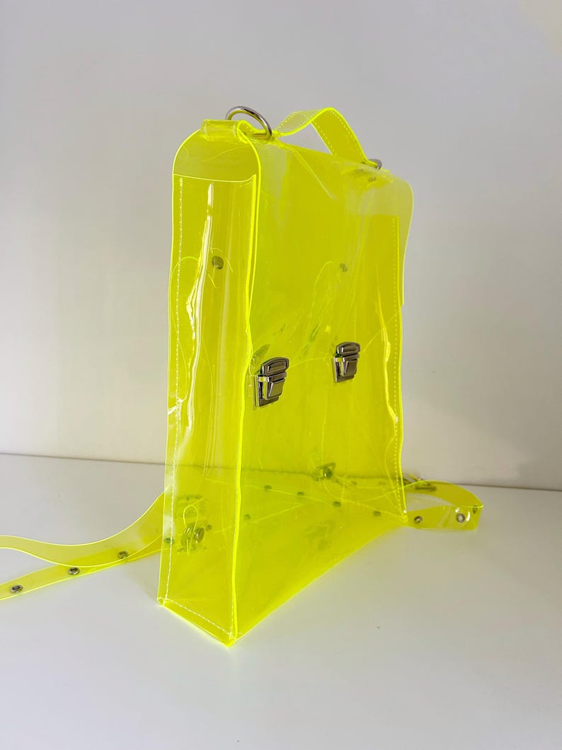 May include: A neon yellow transparent backpack with silver-colored metal clasps and hardware. The bag has adjustable straps and a top handle. The clear material allows visibility of the contents inside. The backpack is designed with a rectangular shape.