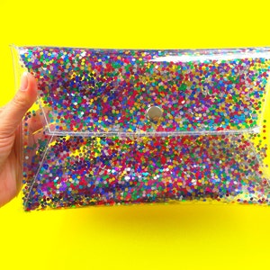 Può includere: Una pochette trasparente in plastica con chiusura a bottone argentata. La borsa è piena di coriandoli colorati.