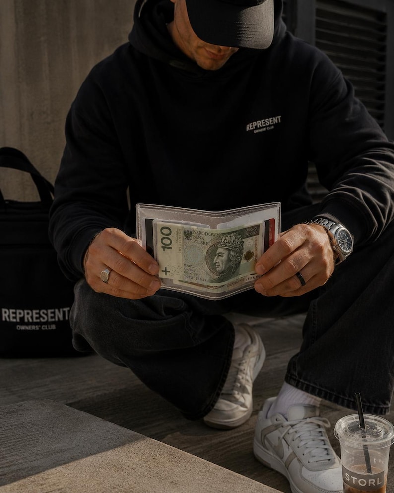 May include: A clear, transparent plastic wallet holding a 100 Polish Zloty banknote featuring a portrait of King Wladyslaw II Jagiello. The wallet is held open, displaying the currency clearly within its rectangular, minimalist frame.