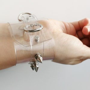 May include: A clear plastic wrist cuff with silver metal accents. The cuff has a buckle closure and three silver spikes.