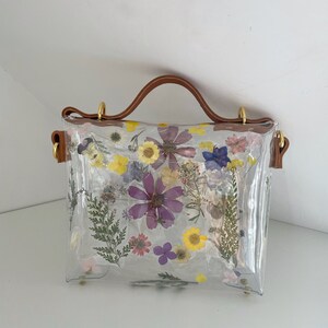 May include: A clear plastic handbag with a brown leather handle and a floral design. The flowers are pressed and dried, and they are arranged in a random pattern. The bag has a gold metal clasp.