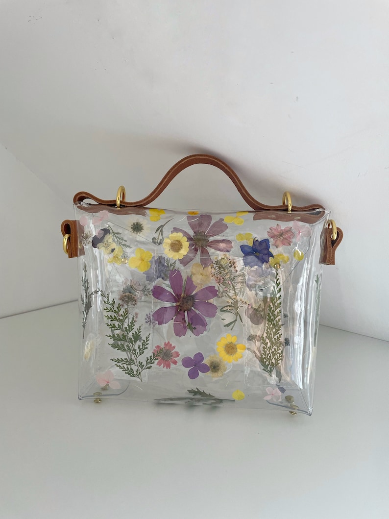 May include: A clear plastic handbag with a brown leather handle and a floral design. The flowers are pressed and dried, and they are arranged in a random pattern. The bag has a gold metal clasp.