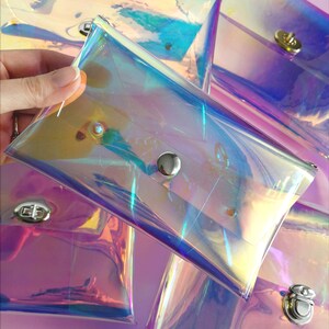 May include: Five iridescent clear plastic pouches with silver snap closures. The pouches are stacked on top of each other and are arranged in a fan shape. The pouches are on a pink background.
