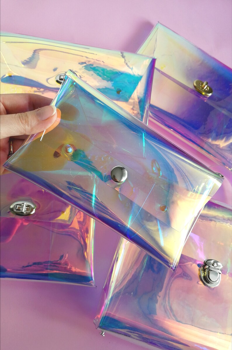 May include: Five iridescent clear plastic pouches with silver snap closures. The pouches are stacked on top of each other and are arranged in a fan shape. The pouches are on a pink background.