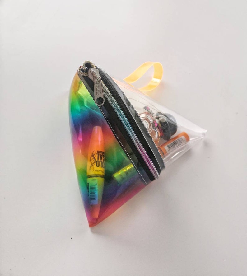 May include: A clear, triangular zippered pouch with a rainbow iridescent design. The pouch is open and contains various items, including a mascara tube, a highlighter, and a small bottle of perfume.