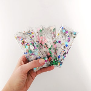 May include: Three clear plastic wallets with different colored sequins and stars inside. The wallets are held in a hand.