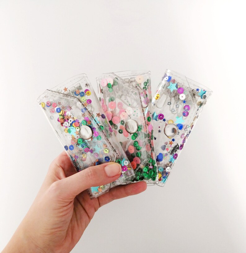 May include: Three clear plastic wallets with different colored sequins and stars inside. The wallets are held in a hand.