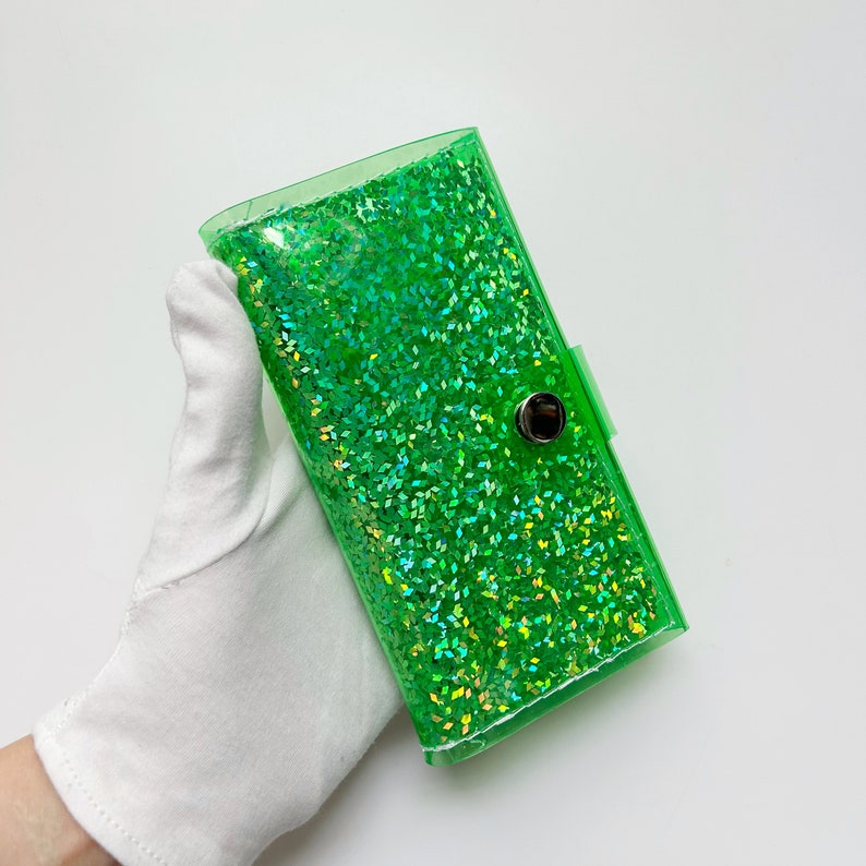 May include: A rectangular, transparent green wallet filled with sparkling green glitter. The wallet has a snap closure and is held by a gloved hand. The glitter catches the light, creating a shimmering effect.