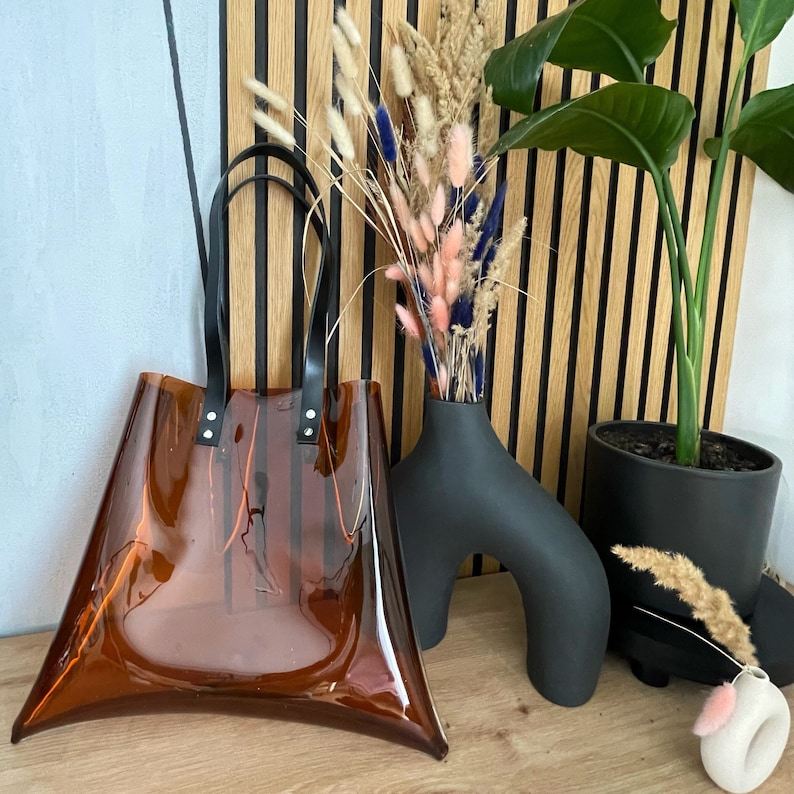 Puede incluir: Un bolso tote marr&oacute;n transparente con asas y detalles en negro. El bolso est&aacute; hecho de un material transparente y tiene forma trapezoidal. El bolso se exhibe junto a un jarr&oacute;n negro con flores secas y una planta en maceta.