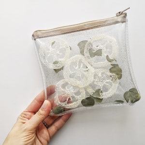 Puede incluir: Una bolsa transparente y brillante con cremallera que contiene varias esponjas de luffa blanquecinas y hojas de eucalipto verdes. La bolsa tiene una cremallera y un ribete beiges.