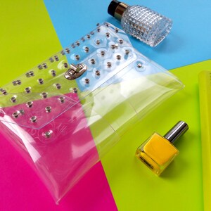 Puede incluir: Un bolso de mano transparente de pl&aacute;stico con tachuelas plateadas y un cierre plateado. El bolso est&aacute; sobre un fondo rosa, verde y azul.