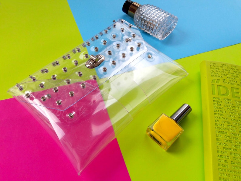 Puede incluir: Un bolso de mano transparente de pl&aacute;stico con tachuelas plateadas y un cierre plateado. El bolso est&aacute; sobre un fondo rosa, verde y azul.