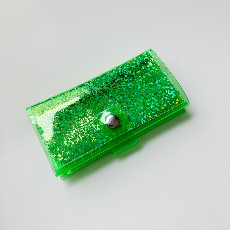 May include: A rectangular, transparent green glitter wallet with a silver snap closure. The wallet is filled with sparkling green glitter, creating a shimmering effect. The wallet is made of a clear, flexible material.