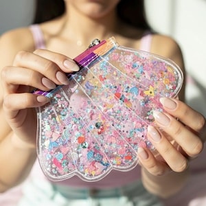 Könnte beinhalten: Eine transparente, muschelförmige Tasche gefüllt mit bunten Konfetti, Sternen und kleinen dekorativen Gegenständen. Die Tasche hat einen Regenbogenreißverschluss und wird von einer Person gehalten. Die Tasche ist mit rosa, blauen, gelben und weißen Dekorationen gefüllt.