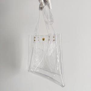 K&ouml;nnte beinhalten: Eine transparente Tragetasche mit rechteckiger Form und trapezf&ouml;rmigem Boden. Die Tasche hat zwei Griffe und wird von einer behandschuhten Hand gehalten. Goldfarbene Nieten und ein Druckknopfverschluss sichern die Oberseite.
