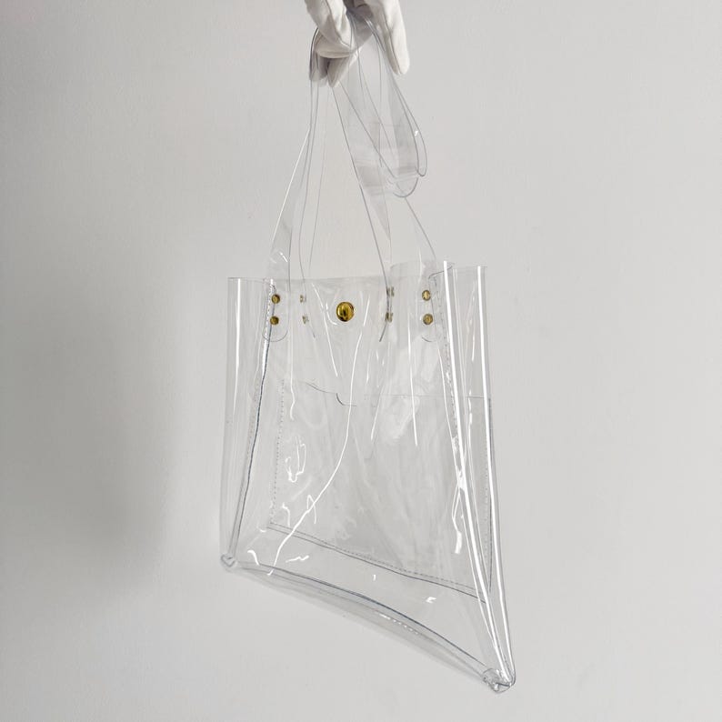 K&ouml;nnte beinhalten: Eine transparente Tragetasche mit rechteckiger Form und trapezf&ouml;rmigem Boden. Die Tasche hat zwei Griffe und wird von einer behandschuhten Hand gehalten. Goldfarbene Nieten und ein Druckknopfverschluss sichern die Oberseite.