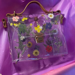 May include: A clear plastic handbag with a brown leather handle and a gold clasp. The bag is decorated with pressed flowers in various colors, including purple, yellow, and red.