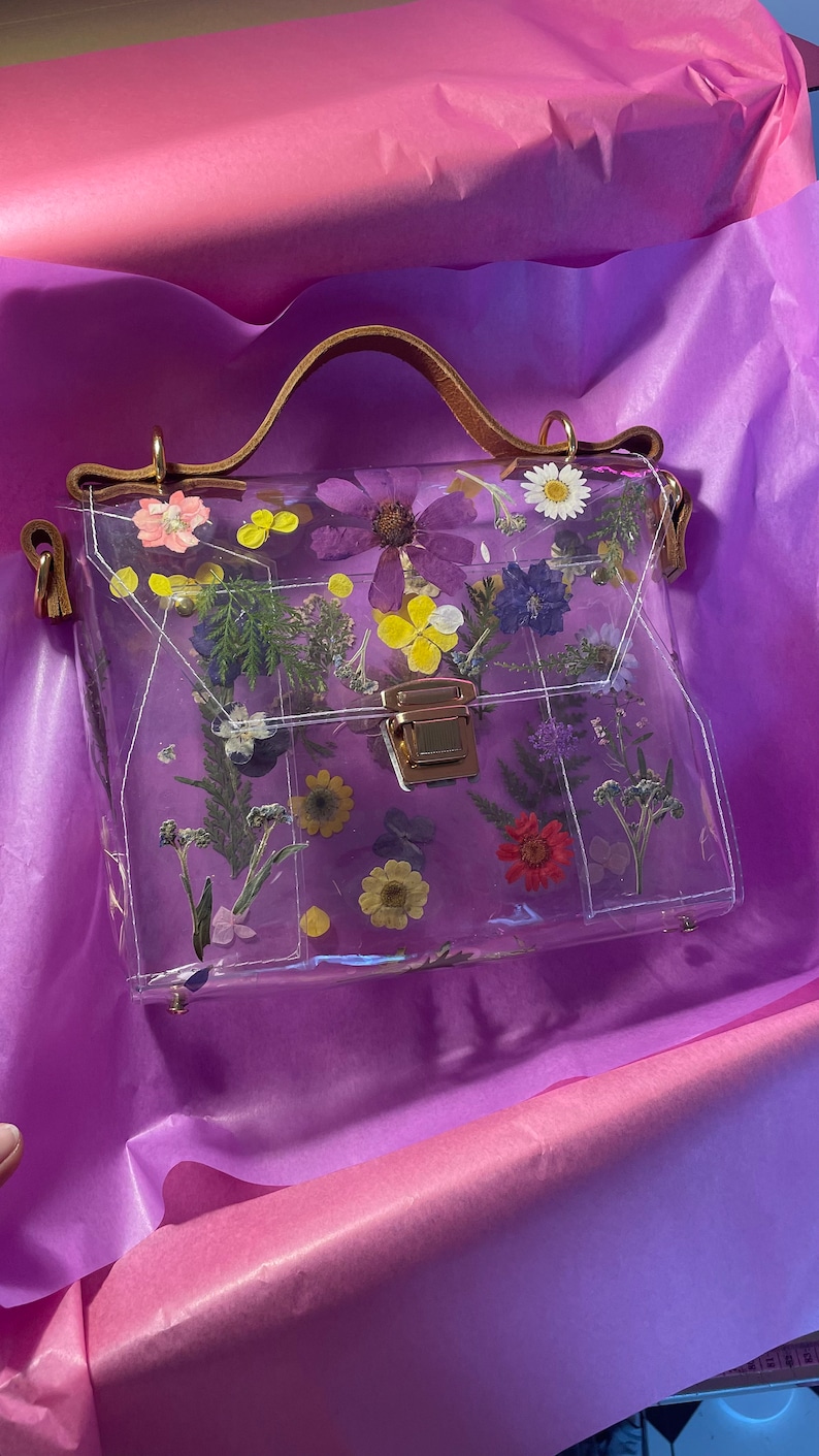 May include: A clear plastic handbag with a brown leather handle and a gold clasp. The bag is decorated with pressed flowers in various colors, including purple, yellow, and red.