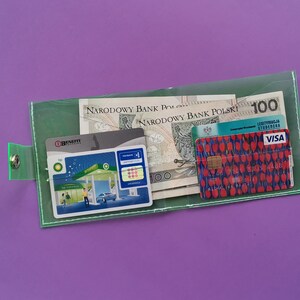 Pu&ograve; includere: Un portafoglio verde trasparente con chiusura a scatto, contenente banconote polacche, carte di credito e una carta fedelt&agrave; della stazione di servizio BP. Il portafoglio &egrave; su uno sfondo viola. La carta di credito ha un motivo floreale rosso.