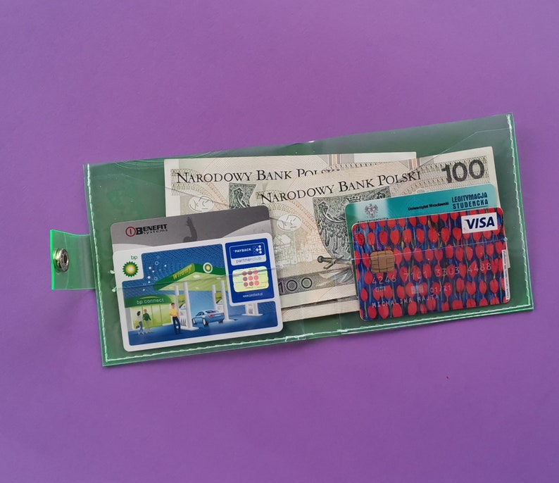 Pu&ograve; includere: Un portafoglio verde trasparente con chiusura a scatto, contenente banconote polacche, carte di credito e una carta fedelt&agrave; della stazione di servizio BP. Il portafoglio &egrave; su uno sfondo viola. La carta di credito ha un motivo floreale rosso.
