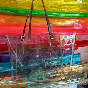 Puede incluir: Un bolso tote transparente e incoloro de forma rectangular, con un bolsillo frontal y un cierre de bot&oacute;n a presi&oacute;n. El bolso incluye dos correas de cuero marr&oacute;n oscuro sujetas con remaches met&aacute;licos plateados en el borde superior.
