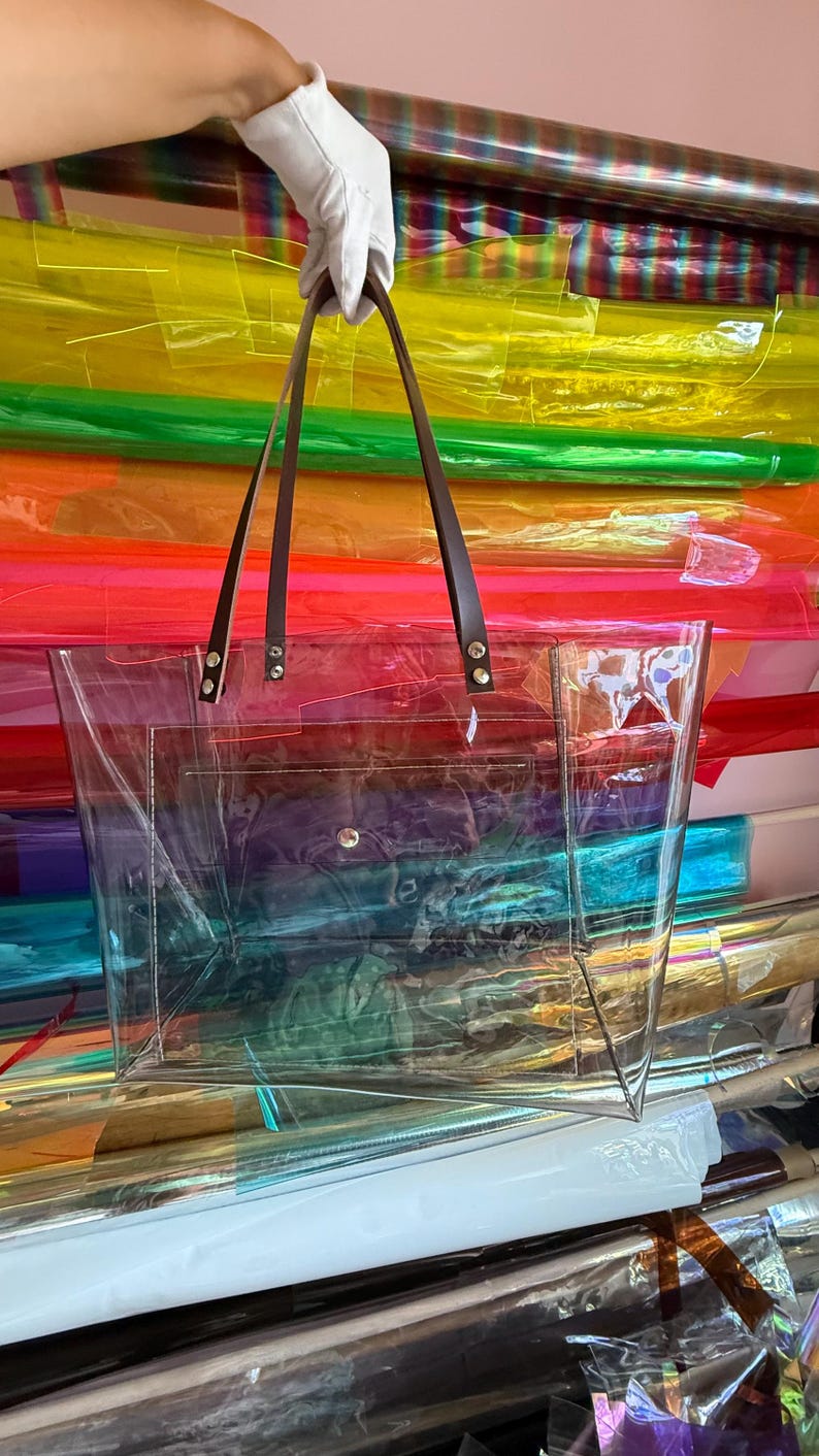 Puede incluir: Un bolso tote transparente e incoloro de forma rectangular, con un bolsillo frontal y un cierre de bot&oacute;n a presi&oacute;n. El bolso incluye dos correas de cuero marr&oacute;n oscuro sujetas con remaches met&aacute;licos plateados en el borde superior.