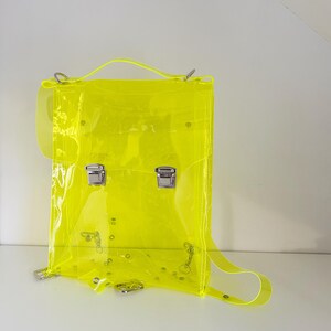 May include: A neon yellow transparent backpack with silver-colored hardware. The bag has a top handle, adjustable shoulder straps, and two front clasps. The backpack is made of a clear, flexible material.