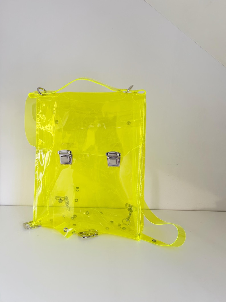 May include: A neon yellow transparent backpack with silver-colored hardware. The bag has a top handle, adjustable shoulder straps, and two front clasps. The backpack is made of a clear, flexible material.
