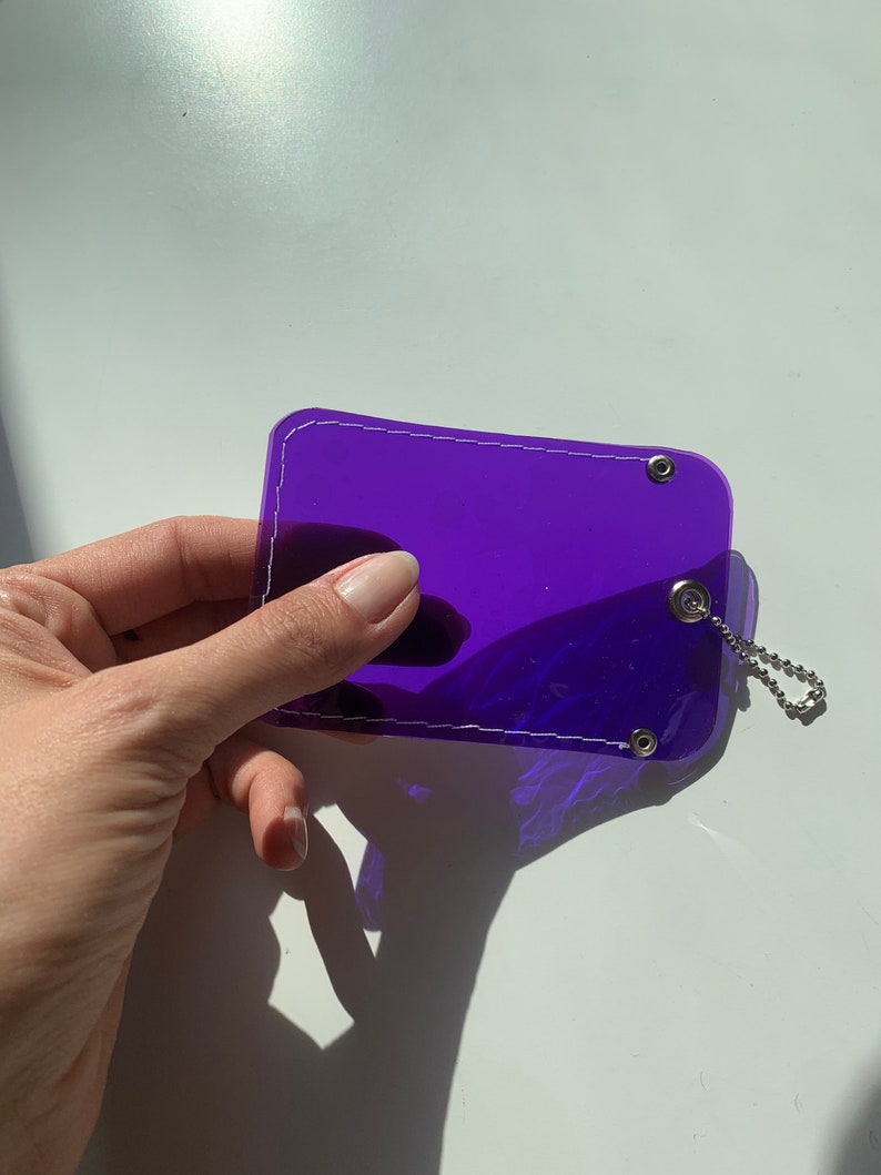 May include: A purple plastic card holder with a white stitched border and silver metal rings. The card holder has a silver chain attached.