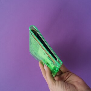 Pu&ograve; includere: Un portafoglio trasparente verde neon con chiusura a scatto, pieno di contanti e carte. Il portafoglio &egrave; tenuto su uno sfondo viola, mettendo in risalto il suo contenuto e il suo design. I bordi del portafoglio sono bordati di verde brillante.