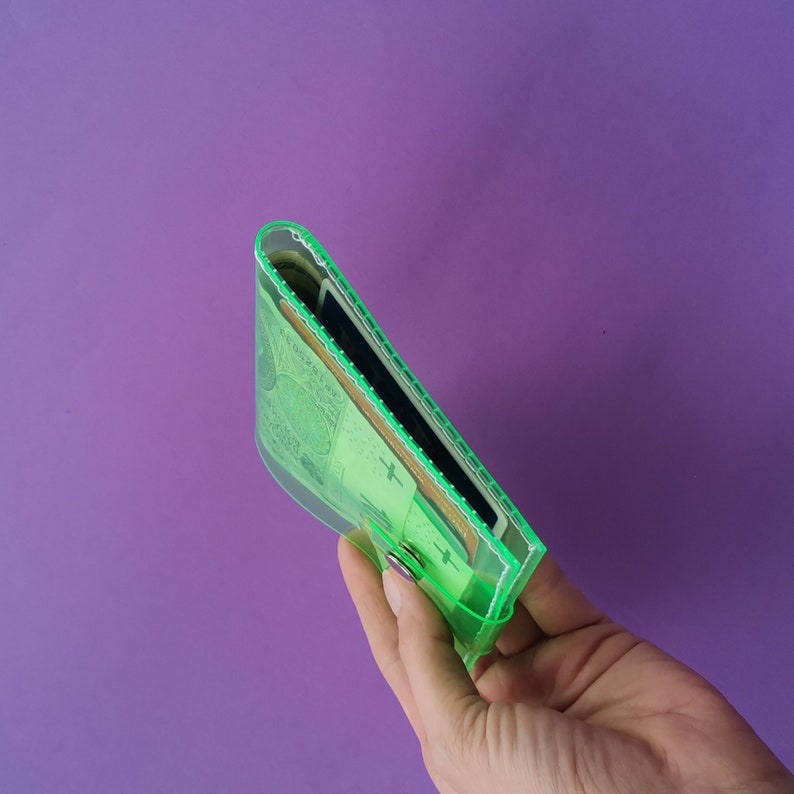 Pu&ograve; includere: Un portafoglio trasparente verde neon con chiusura a scatto, pieno di contanti e carte. Il portafoglio &egrave; tenuto su uno sfondo viola, mettendo in risalto il suo contenuto e il suo design. I bordi del portafoglio sono bordati di verde brillante.