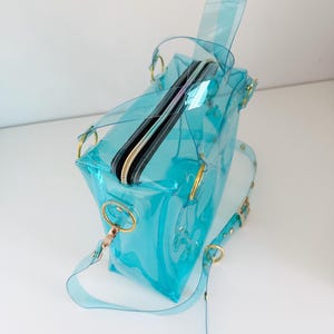 May include: A transparent, turquoise handbag with a zippered top and gold-colored hardware. The bag has a shoulder strap and a top handle. The bag is made of a clear, flexible material.