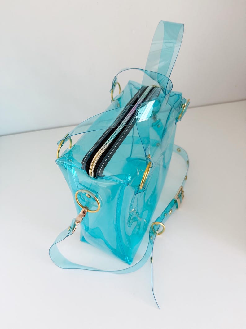May include: A transparent, turquoise handbag with a zippered top and gold-colored hardware. The bag has a shoulder strap and a top handle. The bag is made of a clear, flexible material.