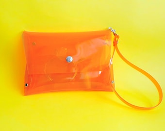 May include: A transparent orange clutch bag with a matching wrist strap. The bag has a rectangular shape with rounded corners and is secured with a silver snap closure. The background is a bright yellow.