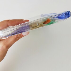 Puede incluir: Una bolsa de pl&aacute;stico transparente con un forro de tela azul. La bolsa contiene una variedad de flores secas y una peque&ntilde;a mariposa naranja.