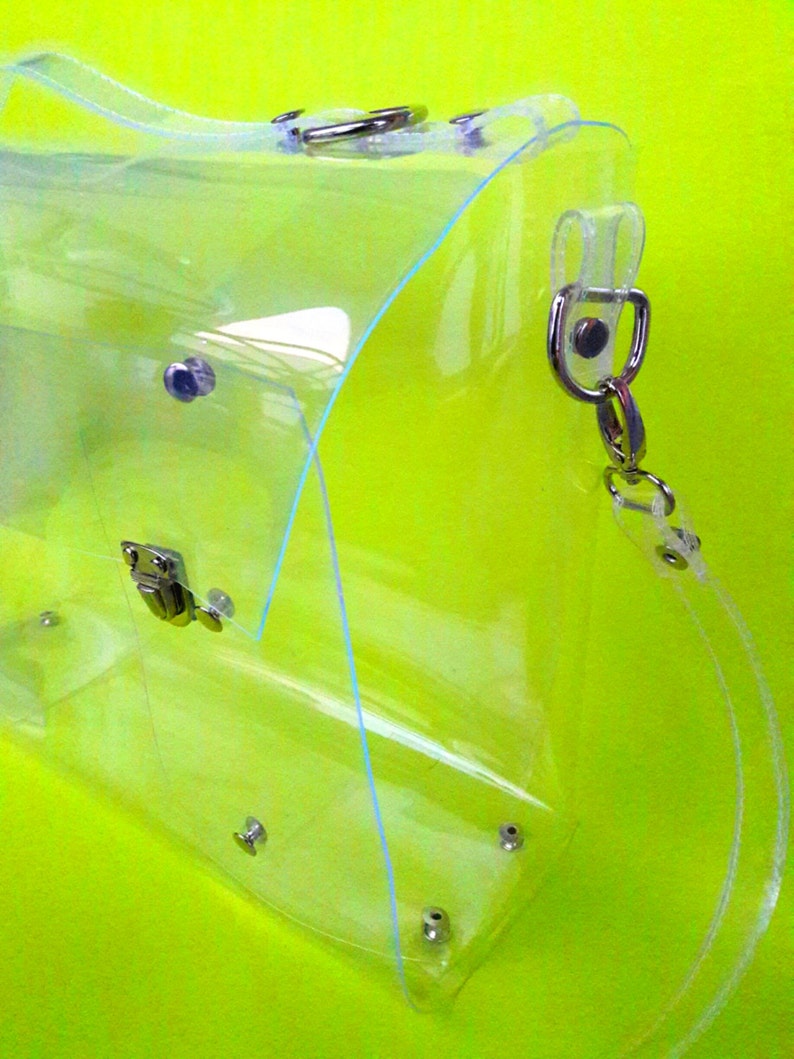 May include: A clear plastic tote bag with a silver chain strap. The bag has a flap closure with a silver buckle.