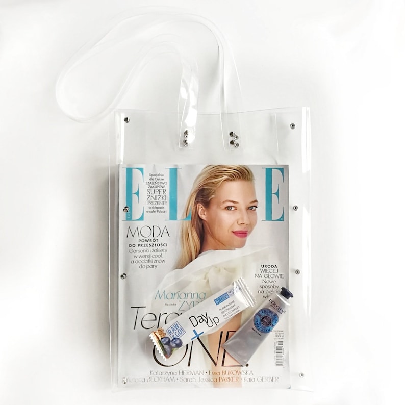 May include: A clear plastic tote bag with a silver zipper and silver studs. The bag contains a magazine, a beauty product, and a snack bar. The magazine is Elle and features a woman on the cover. The beauty product is a tube of hand cream. The snack bar is a blueberry granola bar.