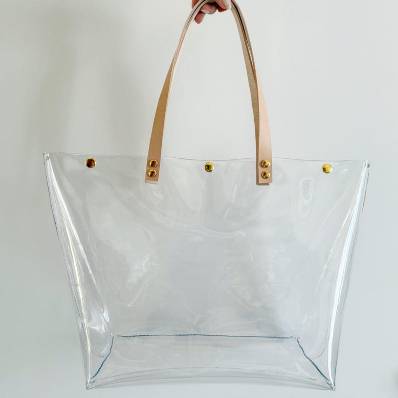 May include: A clear tote bag with tan leather handles and gold-colored rivets. The bag is made of transparent material, allowing visibility of the contents. The handles are attached with gold rivets. The bag is held up by a hand.
