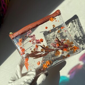 May include: A clear plastic pouch with a brown zipper and a floral design. The pouch is filled with dried flowers and glitter.