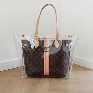 May include: Clear tote bag with brown monogram pattern and orange stripe. Features tan leather handles and trim. The initials "JS" are printed on the orange stripe. Gold-tone hardware accents the bag's design. A stylish and functional accessory.