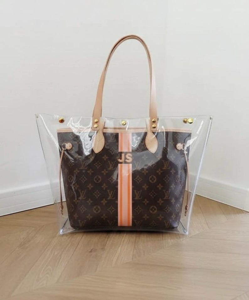 May include: Clear tote bag with brown monogram pattern and orange stripe. Features tan leather handles and trim. The initials "JS" are printed on the orange stripe. Gold-tone hardware accents the bag's design. A stylish and functional accessory.