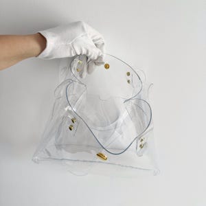 K&ouml;nnte beinhalten: Eine transparente Handtasche mit goldfarbenen Beschl&auml;gen. Die Tasche hat einen einzigartigen, geschwungenen Griff und wird von einer behandschuhten Hand gehalten. Das Design der Tasche ist durch das transparente Material sichtbar.