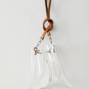 May include: A clear transparent bucket bag featuring a tan leather strap with a knotted design. The bag includes silver-toned metal hardware, a white rope drawstring closure with metal grommets, and a small white tassel at the bottom corner.