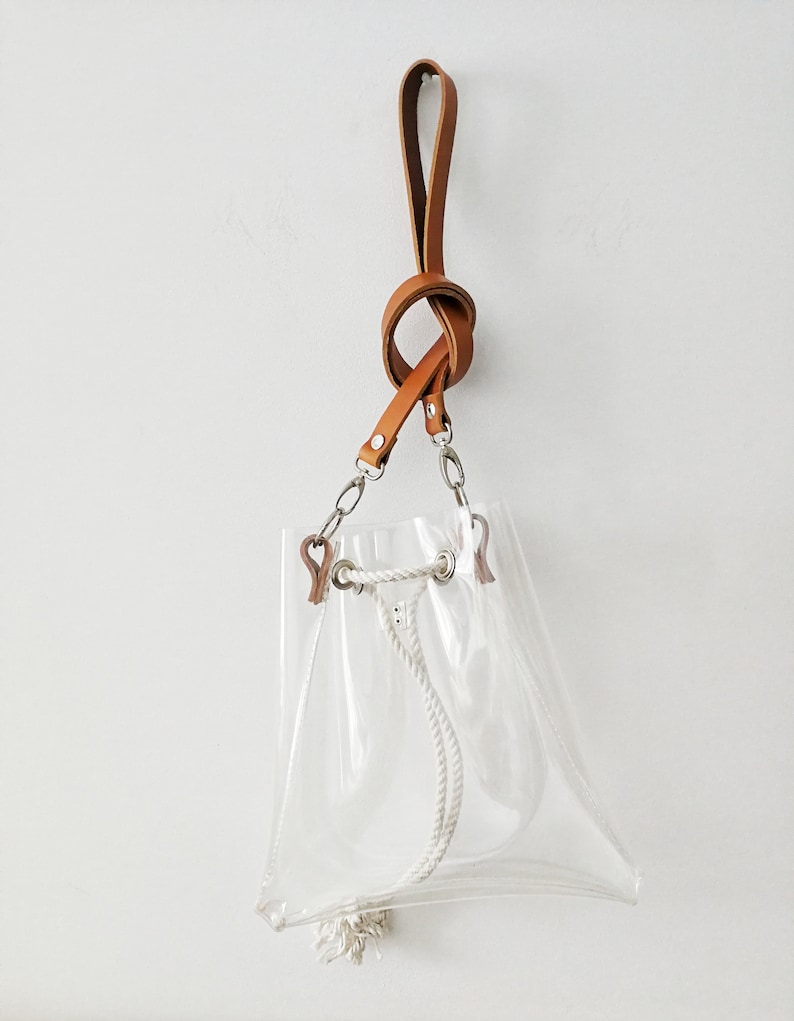 May include: A clear transparent bucket bag featuring a tan leather strap with a knotted design. The bag includes silver-toned metal hardware, a white rope drawstring closure with metal grommets, and a small white tassel at the bottom corner.