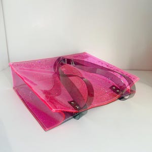 Puede incluir: Un bolso tote rosa transparente con purpurina y asas rosa oscuro. El bolso es rectangular con fondo plano y tiene un aspecto brillante y brillante. Las asas est&aacute;n sujetas con remaches plateados.