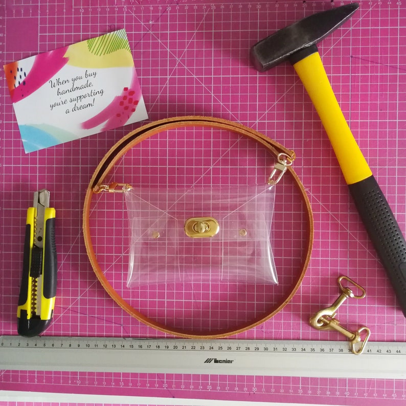 May include: A clear plastic bag with a gold clasp, a brown leather strap, and a metal hook. The bag is on a pink cutting mat with a ruler and a hammer.
