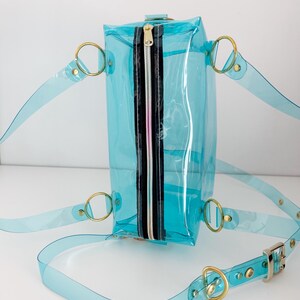 May include: A clear, turquoise-colored backpack with gold-colored metal hardware. The bag has a rectangular shape with a black and silver zipper. The straps are made of the same clear turquoise material as the bag.