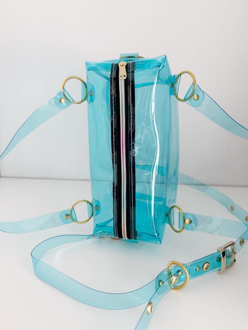 May include: A clear, turquoise-colored backpack with gold-colored metal hardware. The bag has a rectangular shape with a black and silver zipper. The straps are made of the same clear turquoise material as the bag.